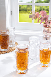 Glamis Cam Fleur De Lis Model Bardak 4'lü Set, açık renkli bir içecekle dolu eşsiz cam bardaklar, doğal bir ortamda pencereden gelen ışıkla birlikte görünmekte. Bu zarif bardak seti, geleneksel tasarımıyla sofistike bir sunum sağlıyor.